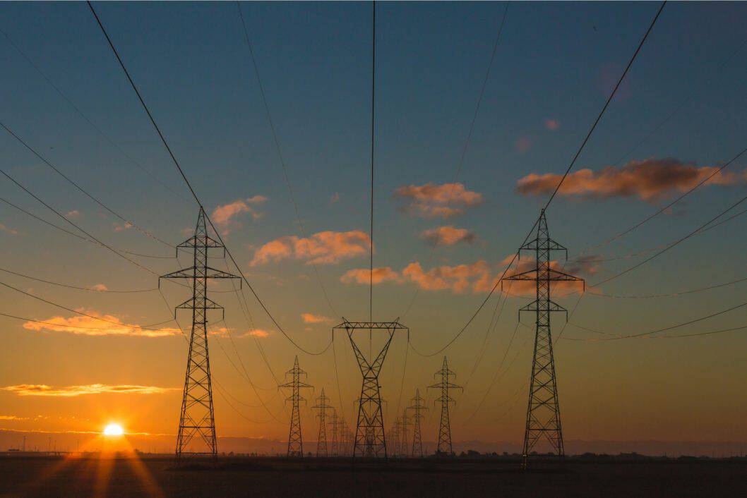 Tall power lines stretch across a flat landscape at sunset, silhouetted against a colorful sky with scattered clouds. No landmarks visible.