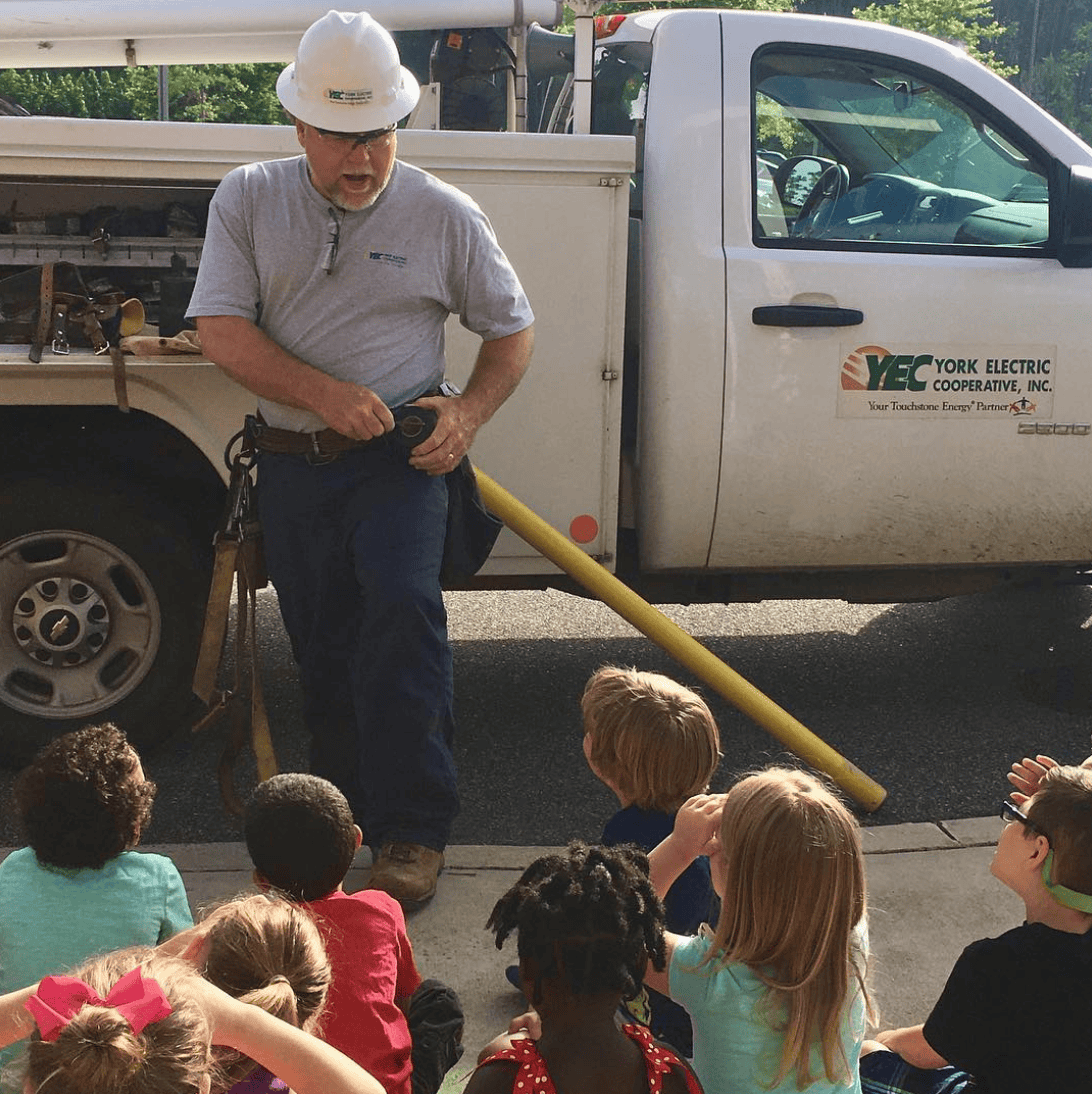 A person in a hard hat talks to children sitting on the ground, next to a York Electric Cooperative truck in sunny weather.