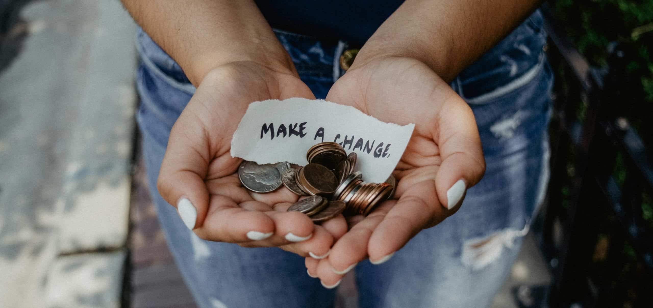 A person holds coins and a note reading "MAKE A CHANGE" in their cupped hands, suggesting a message of charity or positive action.