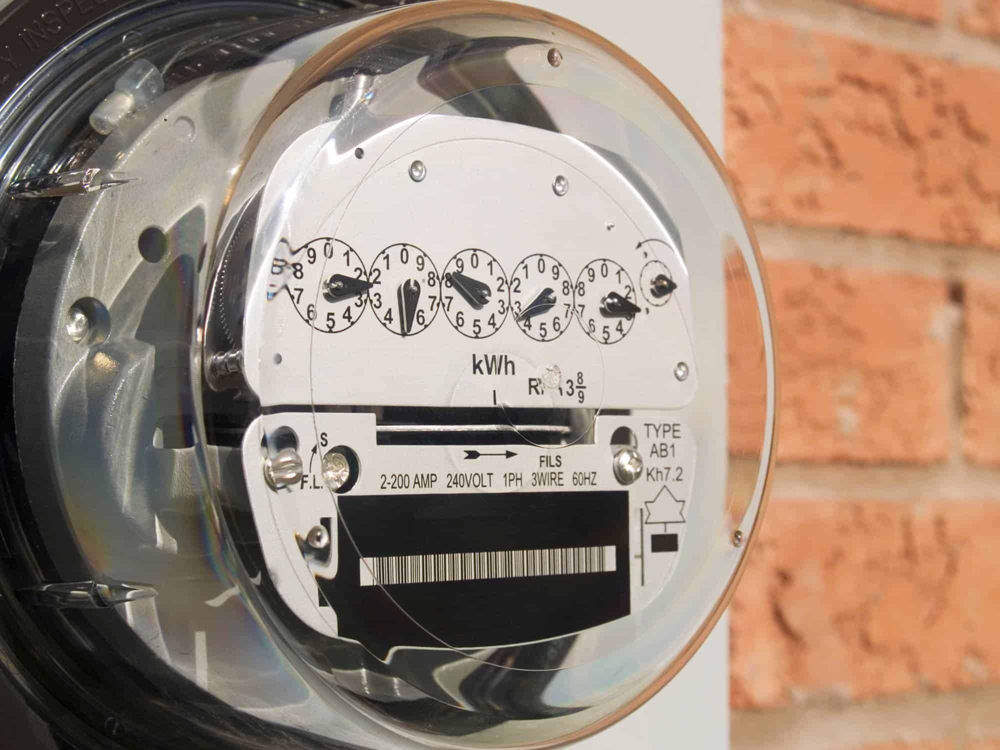 An electric meter measures electricity consumption against a brick wall background, displaying usage in kilowatt-hours with dials and labels.