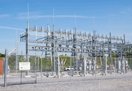 Electrical substation with metal framework, fenced area, sign reading "Alison Green Substation," trees in background under clear blue sky. No people present.