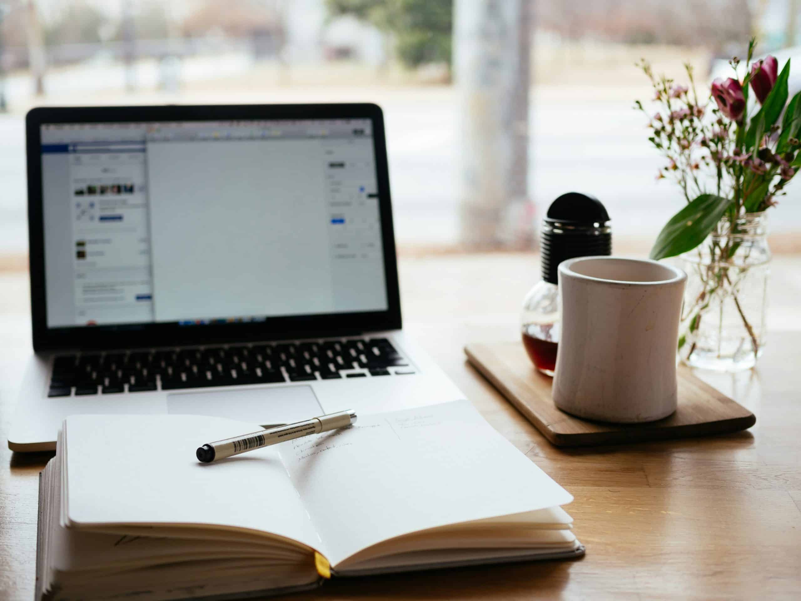 Open laptop, notebook with pen, coffee cup, and flowers on a wooden table near a window with an outdoor view in the background.