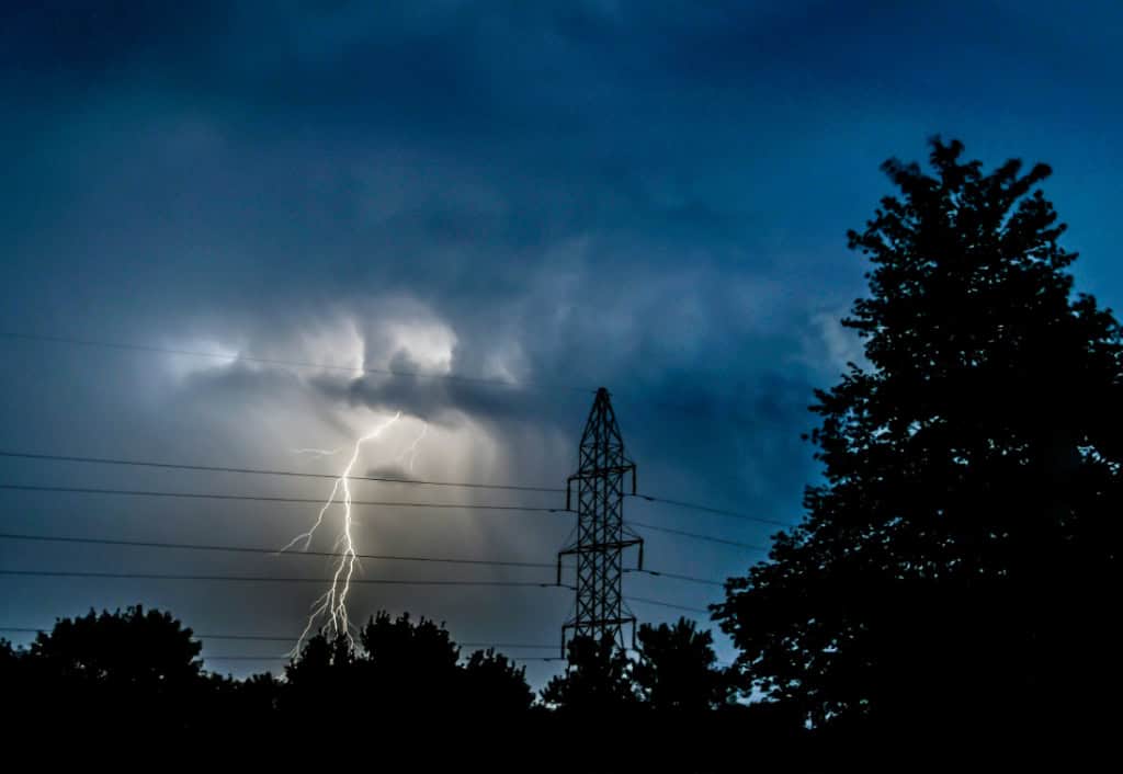 A thunderstorm in a dark sky with a lightning strike behind trees and a transmission tower, with power lines prominently visible.