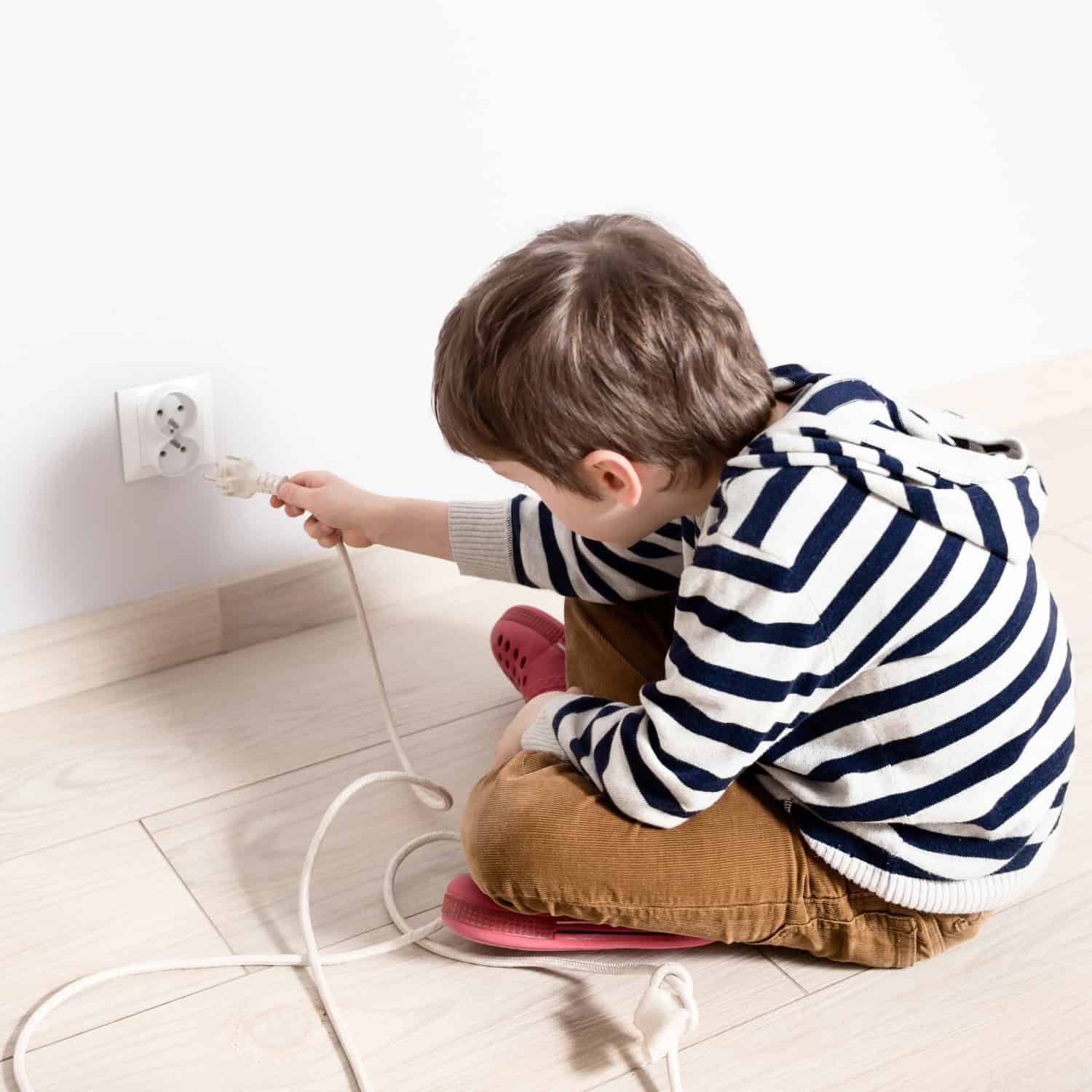 A child wearing a striped hoodie attempts to plug a cord into a wall socket, sitting on a wooden floor.