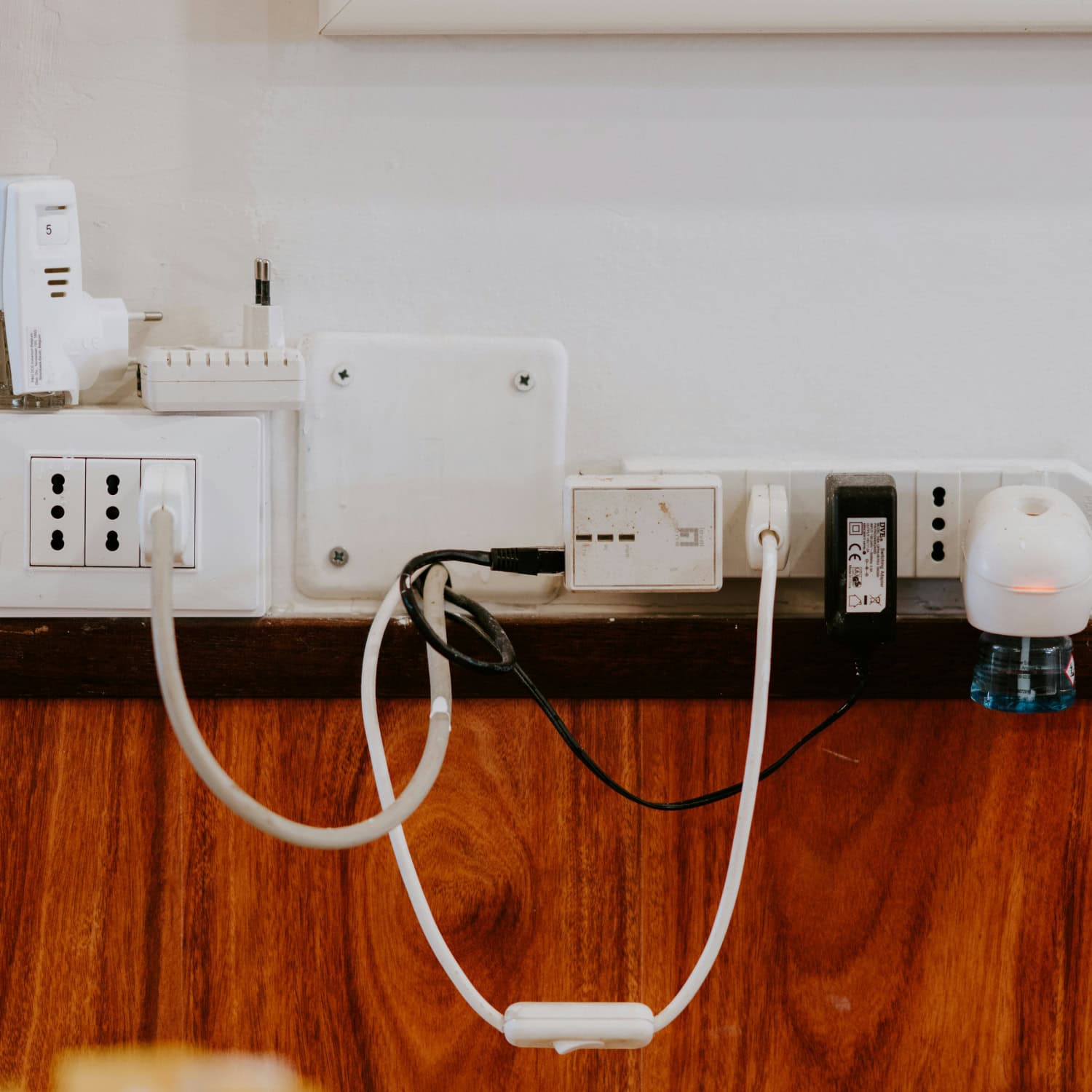 Various electrical plugs and chargers connected to wall sockets on a wooden panel, with a white charger and a room freshener visible.