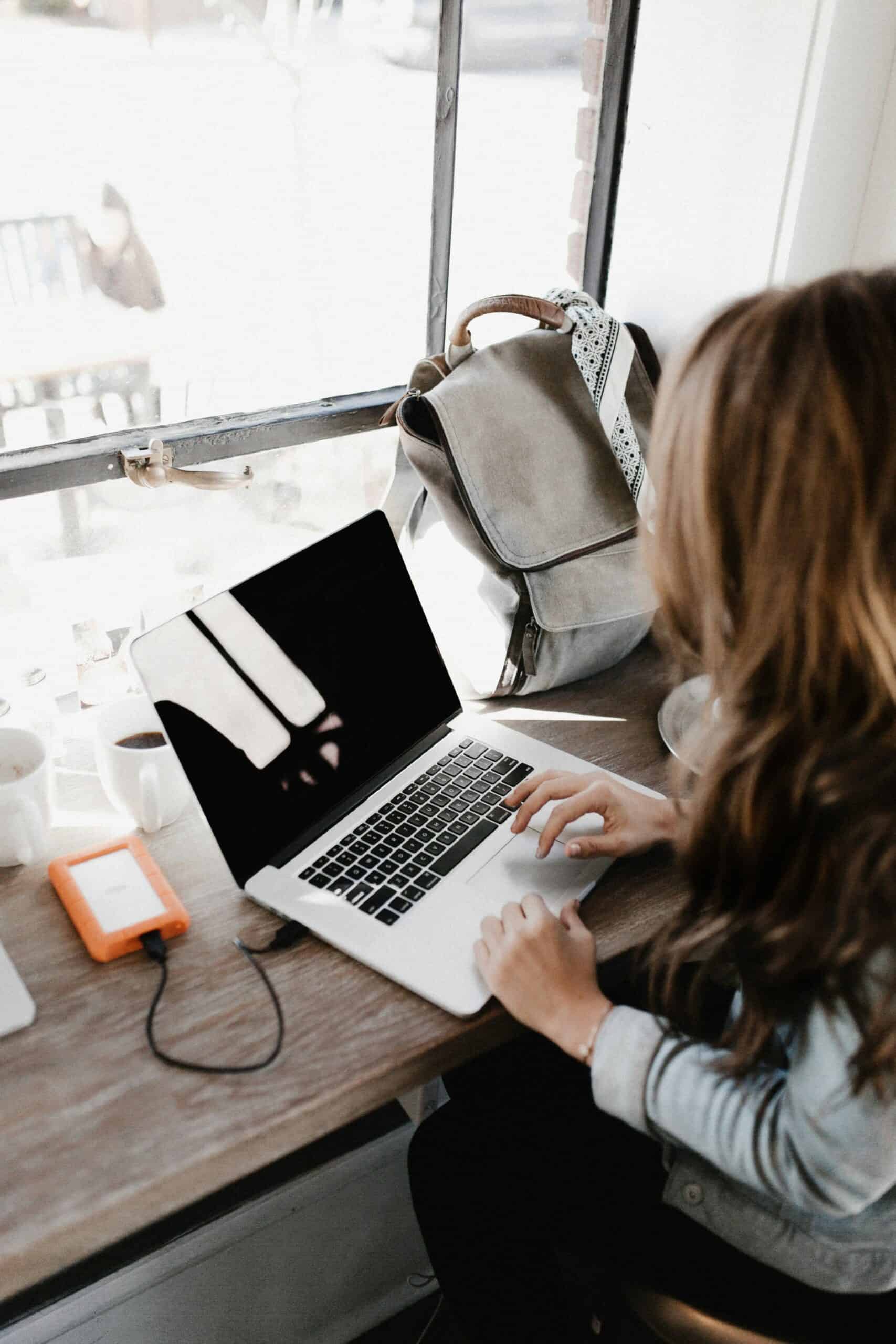 A person using a laptop at a wooden table near a window, with a backpack, coffee cup, and external hard drive present.