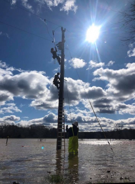 YEC lineworkers climbing a partially submerged utility pole with a crew member in hip waders standing below.