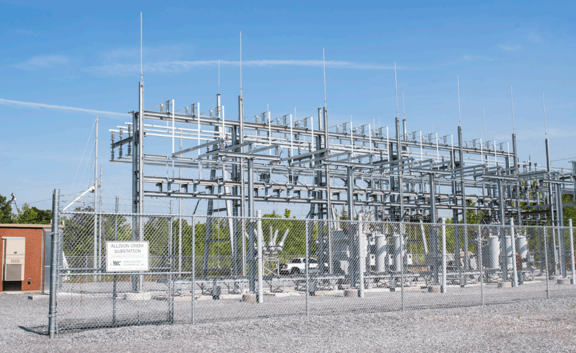 Electrical substation with metal structures and transformers, surrounded by a chain-link fence. A sign is visible, and the sky is clear.