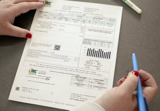 Two hands hold an electricity bill with graphs and details. One hand has red nail polish, and there's a blue pen nearby.