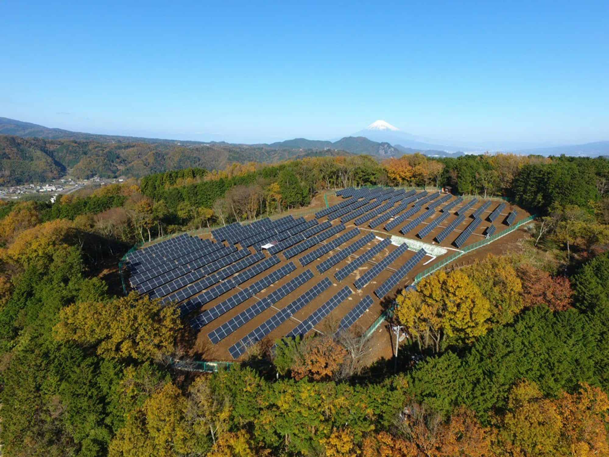 Solar panels in a forested area with Mount Fuji in the background, under a clear blue sky.