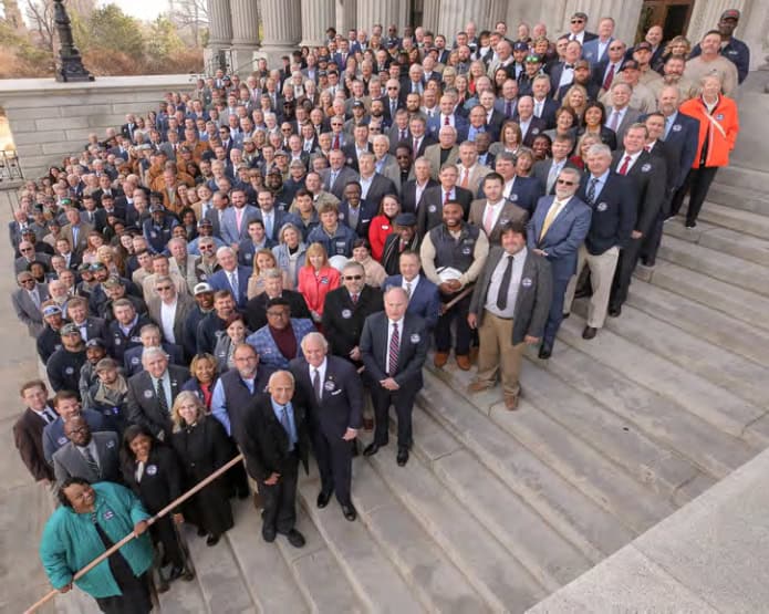 A large group of people stands on the steps of a historical building, possibly a government venue, facing the camera.