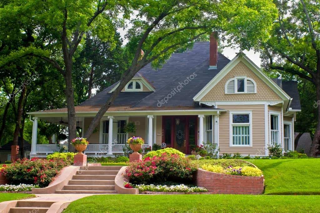 A charming Victorian-style house with a well-manicured lawn, colorful flower beds, large trees, and a welcoming front porch.