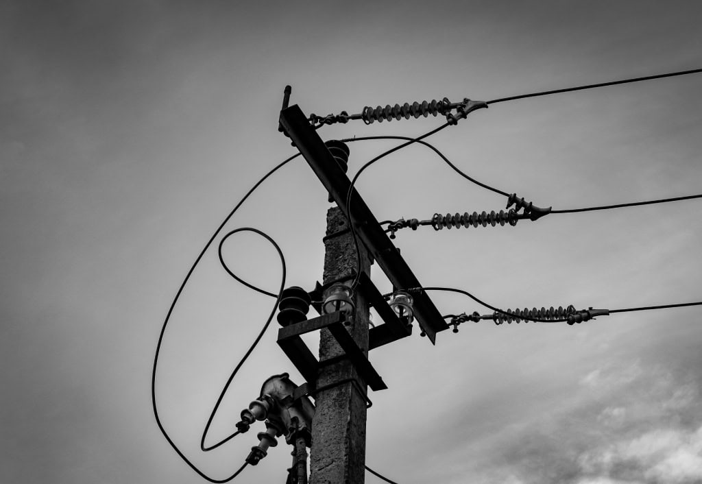 Black and white photo of an electric pole with intricate wires against a cloudy sky, emphasizing industrial elements and contrasting textures.