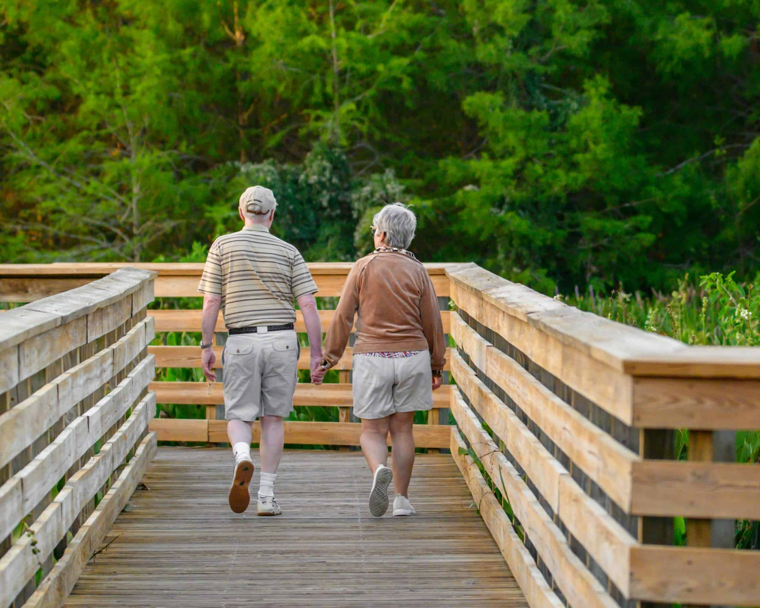 Two people walk hand in hand along a wooden boardwalk surrounded by lush greenery, enjoying a peaceful moment in nature.