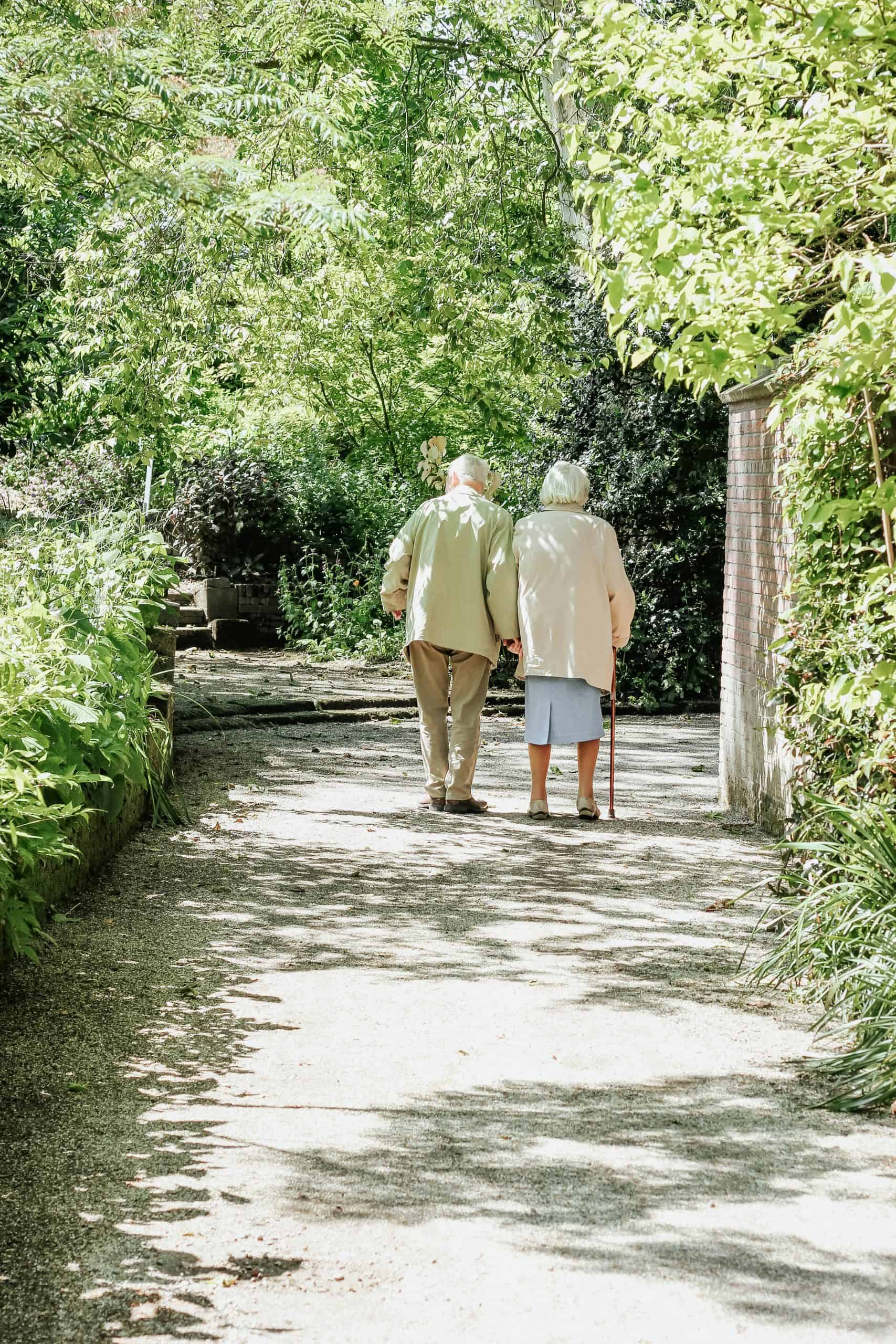 Two people walk down a lush, sunlit garden path, surrounded by green foliage and shaded by trees.