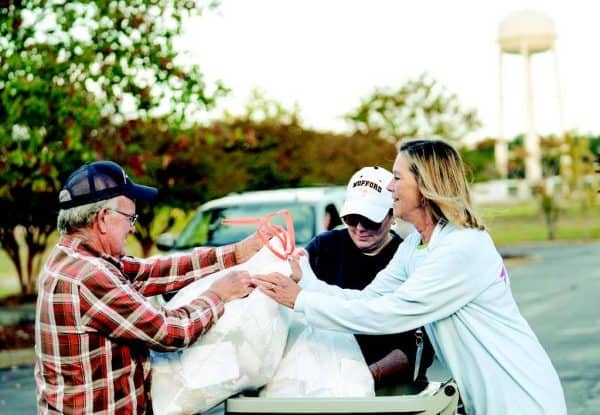 Three people work together outdoors, handling bags by a cart. Trees and a water tower are visible in the background.