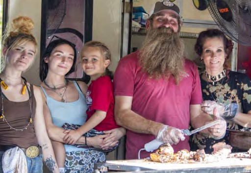 A group of people, including a child, smiles by a food stand. The person in the middle wears a cap and has a large beard.