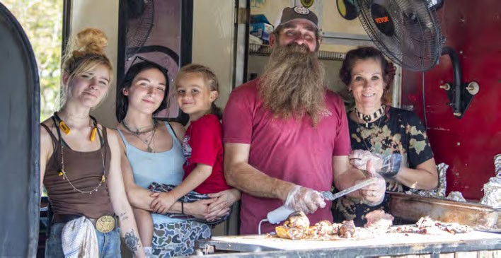 A group of four people and a child smiling, gathered around a food stand or grill with cooked food displayed in front.