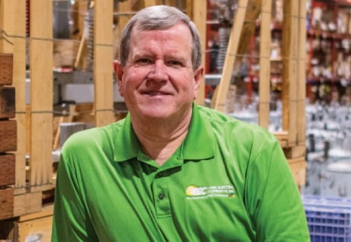 A person in a green collared shirt stands inside a warehouse filled with wooden crates, shelving units, and various stored goods.