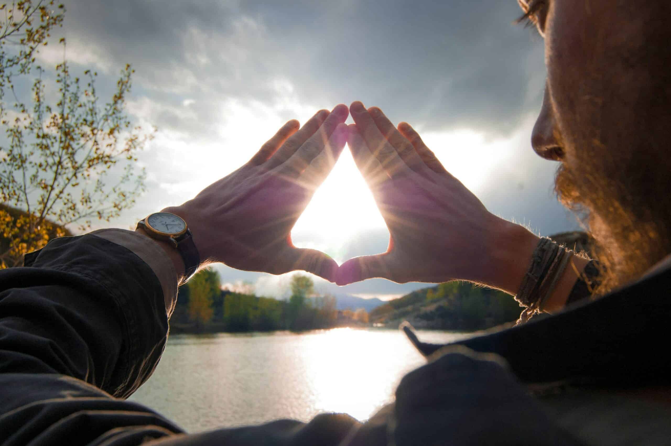 A person frames the setting sun with their hands, standing near a serene lake surrounded by trees, creating a moment of natural beauty.