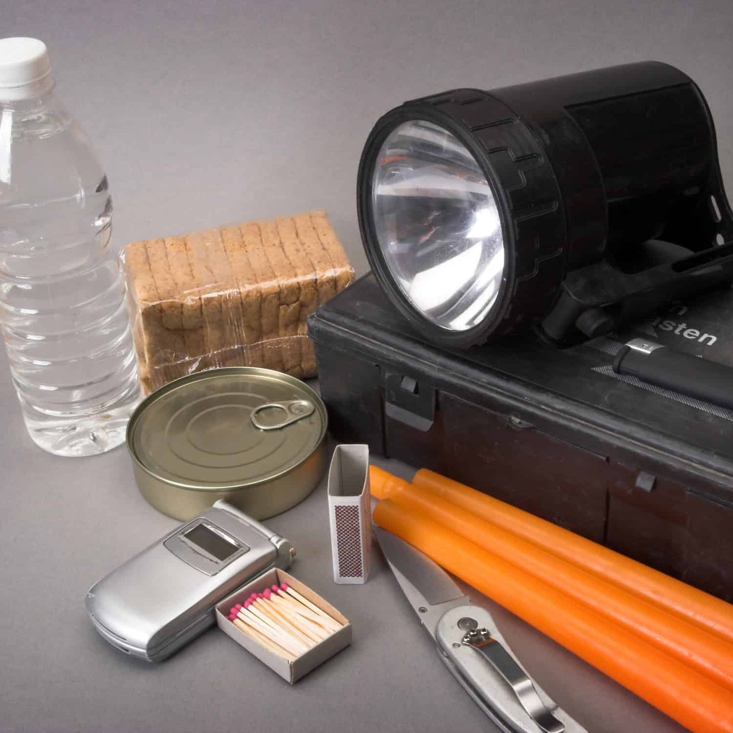 Emergency kit essentials including a flashlight, water bottle, biscuits, canned food, matchbox, candles, multi-tool, phone, and batteries on a gray surface.