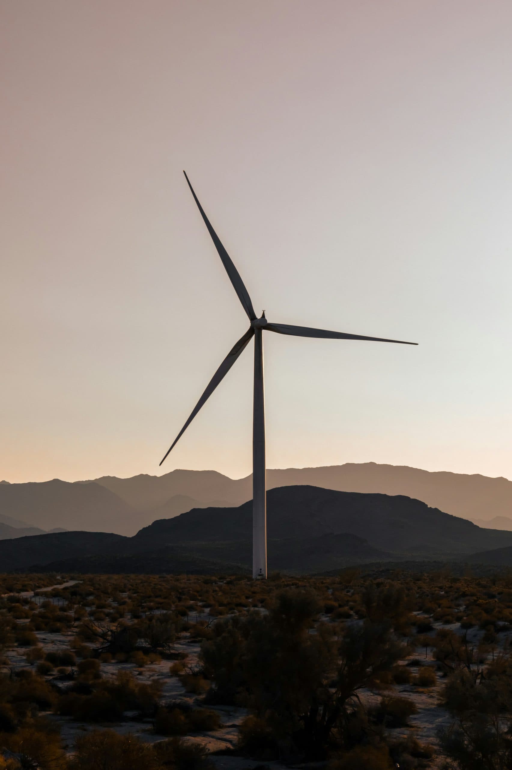 A large wind turbine stands against a mountainous backdrop at sunset, with sparse desert vegetation in the foreground. The sky is softly lit.