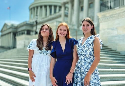 Three people standing in front of the United States Capitol building, smiling, on a sunny day with clear skies.